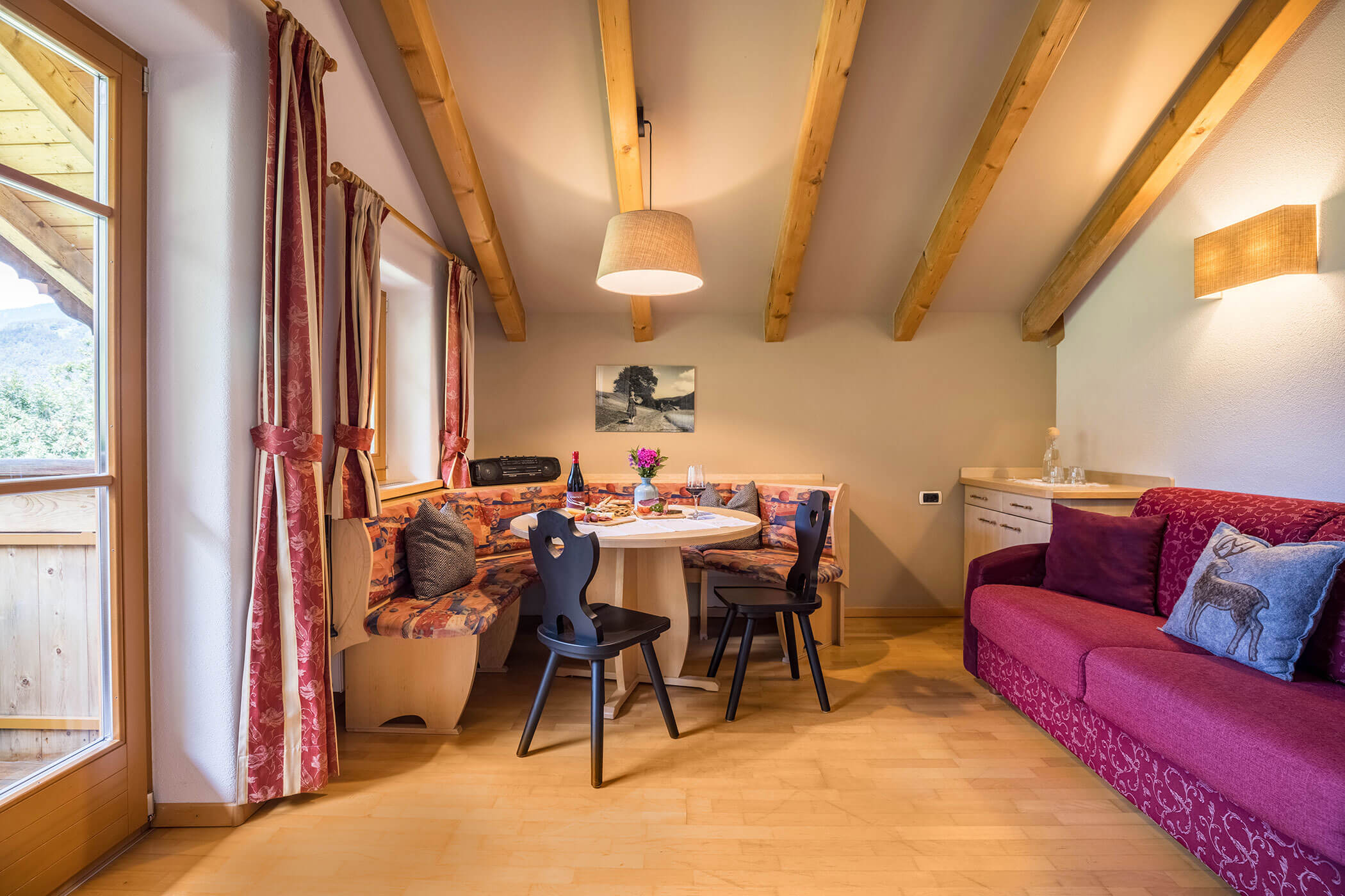 Wohn-Esszimmer mit rotem Sofa, Eckbank, Tisch und zwei Stühlen - Oberplunerhof